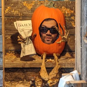 The background is a wooden outhouse frame with a wooden toilet seat. Andrew Cohen's, who has white skin, has his face framed with an orange pumpkin that has arms/hands and legs/feet made of the stems of pumpkins. In the right hand of the pumpkin, is being gripped hard while the left hand has it out in a gesture of "You caught me in the outhouse going to the bathroom"! He is wearing black sunglasses and has a short black beard. He is a closed mouth smile.