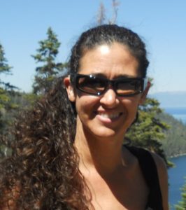 The background has blue skies and green pine trees with Brenda Barrett-Rodriguez standing in front of them. The image shows her from the head to her chest with light tan skin. She has curly dark brown and black hair that is in a side ponytail. She is wearing black framed sunglasses, a smile showing teeth, and is wearing a black tank top.