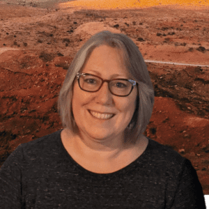 The background has orange, red, and tan rocks with various desert green bushes with Laura Ellington-Pierre standing in front of it. The image shows her from the head to her chest with white skin. She has dark gray and white hair at chin level. She is wearing red framed glasses with a black t-shirt. She has a smile showing teeth