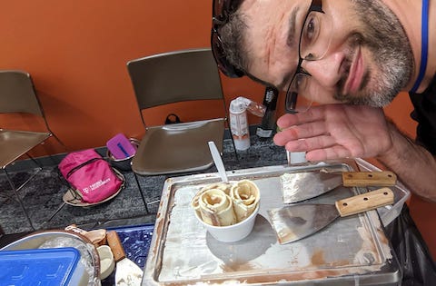 The Rolling Ice Cream [Image Description: The background has an orange wall, several brown plastic and metal chairs, and gray carpet. On the gray carpet, it has various small string backpacks, hats, and a roll of paper towels. The photograph shows a close shot of Andrew Cohen near the stone cold marble slab machine that is empty with 2 spatulas on it, and then a cup of vanilla ice cream that has been rolled. Andrew has sunglasses on his head, black framed glasses, and then has a closed smile while gesturing with his right hand plam up towards the cup of ice cream.