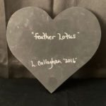 Image Description: [The background is a dark gray color with a black floor. The object in the photograph is in the shape of a heart. The heart is all black with the words “feather Lotus” and “L. Callaghan 2016” in silver.]