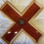 Image Description:[The background is a white tablecloth with a cross made of hard epoxy mixed with color pigmentation made by Steven Peterson. The frame is a light tan color with a dark brown glossy color for the cross. It has a dark green color that looks as though something has “splashed” on it in the middle. The cross does allow light to shine through the dark brown glossy section.]