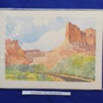 Image Description:[The background is a royal blue tablecloth with a painting made by Tom Posedly of Highway 144, Colorado. It has a blue sky with various white clouds, several rock formations in light, medium, and dark brown, green leafed trees with brown trunks, and a black top highway going through the trees. The words “HIGHWAY 144, COLORADO” are on the bottom left while on the bottom right has “POSEDLY” with both in black.]