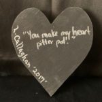 Image Description: [The background is a dark gray color with a black floor. The object in the photograph is in the shape of a heart. The heart is all black with the words “You make my heart pitter pat.” and “L. Callaghan 2017” in silver.]