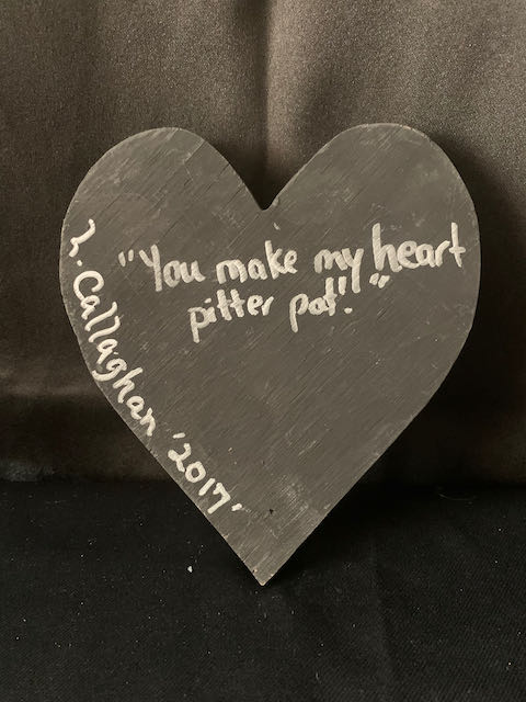 Image Description: [The background is a dark gray color with a black floor. The object in the photograph is in the shape of a heart. The heart is all black with the words “You make my heart pitter pat.” and “L. Callaghan 2017” in silver.]