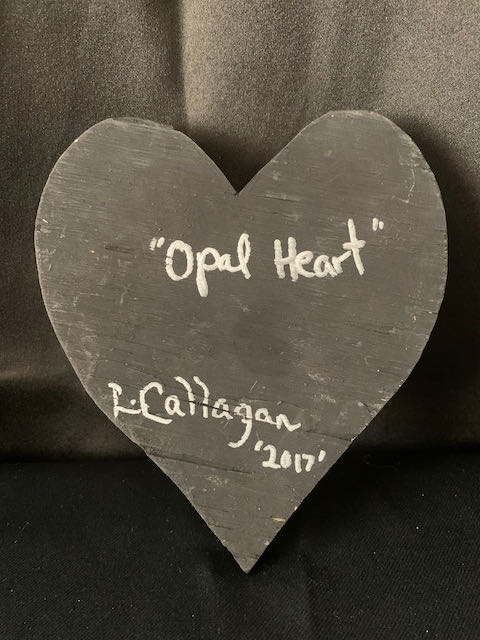 Image Description: [The background is a dark gray color with a black floor. The object in the photograph is in the shape of a heart. The heart is all black with the words “Opal Heart” and “L. Callaghan 2017” in silver.]