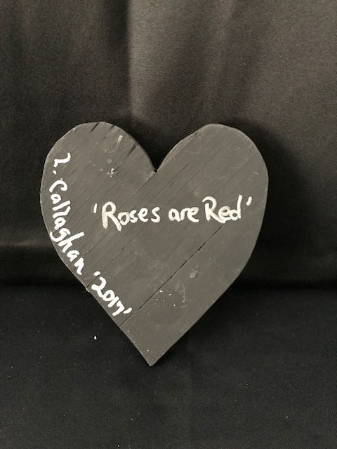 Image Description: [The background is a dark gray color with a black floor. The object in the photograph is in the shape of a heart. The heart is all black with the words “Roses are Red” and “L. Callaghan 2017” in silver.]