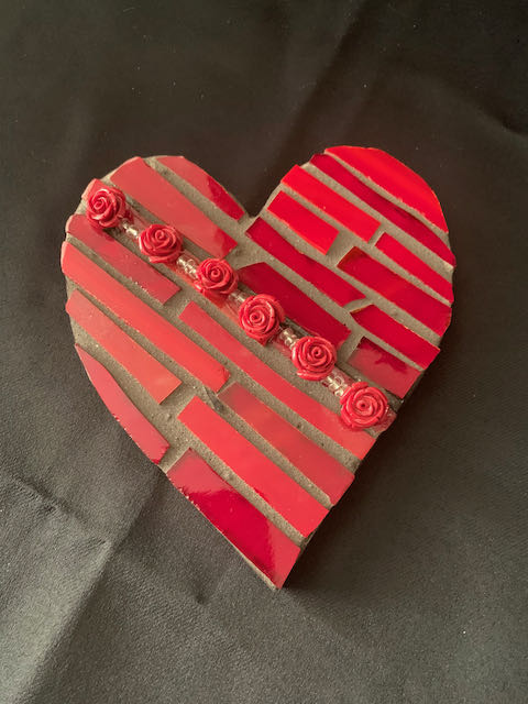 Image Description: [The background is a dark gray color with a black floor. The object in the photograph is in the shape of a heart. The heart has dark gray grout in the background with red ceramic. The red ceramics are all in a rectangle shape that is positioned from the bottom right up to the top left at an angle that looks as though it is stripes. In the middle of the heart, 6 red roses are positioned from the bottom right up to the top left at an angle.]