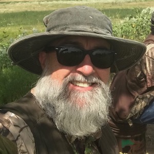 The background shows green grass with hits of the sun shining on it. The close photograph of the white male has on a medium green hunting boonie hat, black framed and lens sunglasses, a white beard down to his chest, a tan long sleeve collared hunting shirt, and a dark green hunting vest.