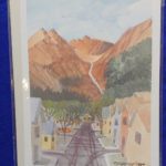 Image Description:[The background is a royal blue tablecloth with a painting made by Tom Posedly of the Telluride, Colorado. It has a white sky with hints of blue, medium and dark brown formations, green leafed trees going down the formation into the neighborhood, rows of house in yellow, blue, and orange with a black top road going down the middle. The word “POSEDLY” written in black at the bottom right.]