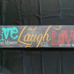 Image Description: [The background is black with a rectangle wooden board with the words “Live EVERY MOMENT”, “Laugh EVERYDAY”, and “Love BEYOND WORDS” from left to right. The word “Live” is a teal blue color, “Laugh” is a mustard yellow color, and “Love” is a red color. The words “EVERY MOMENT”, “EVERYDAY”, and “BEYOND WORDS” are in white.]