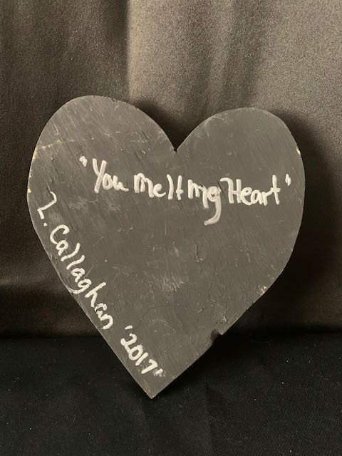 Image Description: [The background is a dark gray color with a black floor. The object in the photograph is in the shape of a heart. The heart has a dark gray grout background with 2 red pieces, 2 pink pieces, and 1 silver metal piece.]