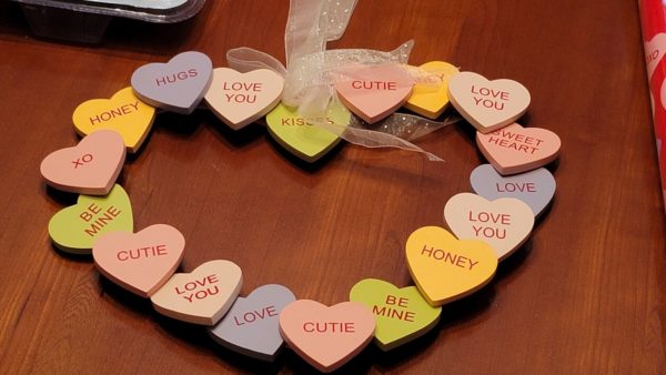 Image Description: [The background is a medium brown color displaying like a desk/table.  The object in the photograph is in the shape of a heart in a wreath style. There are 18 individual miniature hearts connected on a wreath with various texts and colors in them.  All texts are uppercased in pink color with each saying - ‘CUTIE, HONEY, LOVE YOU, XO, SWEETHEART, LOVE, BE MINE and HUGS’.  Colors of each heart randomly in medium pink, light pink, yellow, lime green, and lavender/purple.  On top of the heart is a clear, transparent white bow with glitters.  The back is solid blank with a ring to support the individual, connected hearts.]