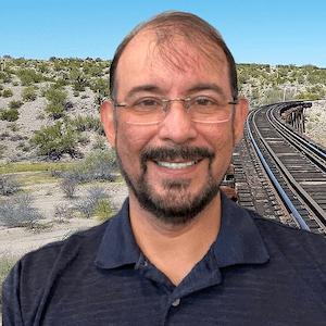 The background has a clear blue sky with various hues of green desert bushes growing in desert sand. On the right side, a set of brown wooden sleepers with metal rails for a train track is seen close, and then at a distance. Pablo Felix is seen from the top of his head to his chest. He has light tan skin with short black hair, silver framed glasses, a black goatee, and then a dark blue polo t-shirt. Pablo is smiling in the photograph with teeth.