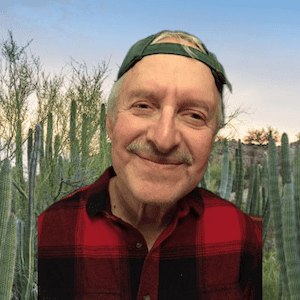 The background shows a light blue and white sky with the sun setting, and then green Organ Pipe Cacti all around. The photograph shows a close headshot of a white man from his head to his shoulders. Martin Greenburg has on a backwards green ball cap with several strands of about 5 inches of white hair seen sticking out from the sides and front. Martin has a white mustach with a closed smile. He is wearing a red and black checkered flannel long sleeve collared shirt.