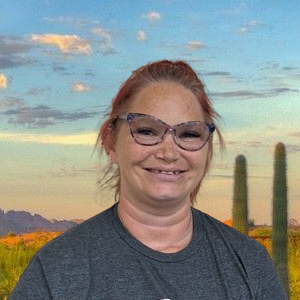 The background has a light blue sky with hues of purple and white, purple looking mountains, and orange sand with two Saguaro cacti on the right. The image shows a white female from the top of her head to her shoulders. The individual's name is DeDe Fisher. She has her hair pulled back in a ponytail with hues of purple and pink in her hair, "cat eye" shaped multicolored framed glasses, a smile showing teeth, and a dark gray t-shirt.